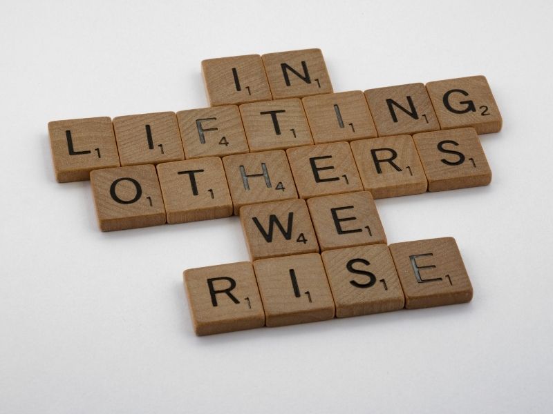 Scrabble tiles spelling "In lifting others we rise," emphasizing supporting others to overcome challenges.