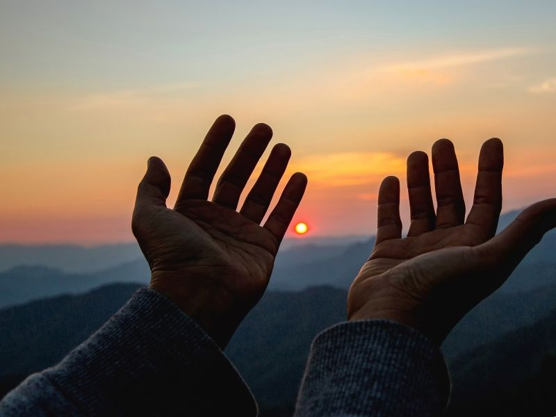 Hands reaching toward a sunset over mountains, illustrating faith, hope, and spiritual resilience.