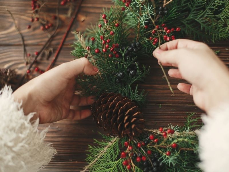 Hands arranging an Advent wreath, with pine cones and red berries, highlighting the spiritual practice of the Advent wreath devotion.