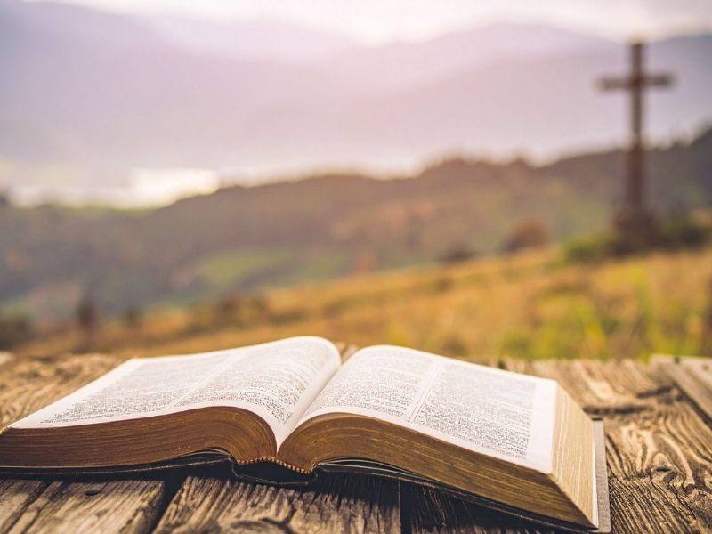 Open Bible on a rustic wooden table with a scenic mountain landscape with the Cross in the background, representing faith and spiritual renewal.