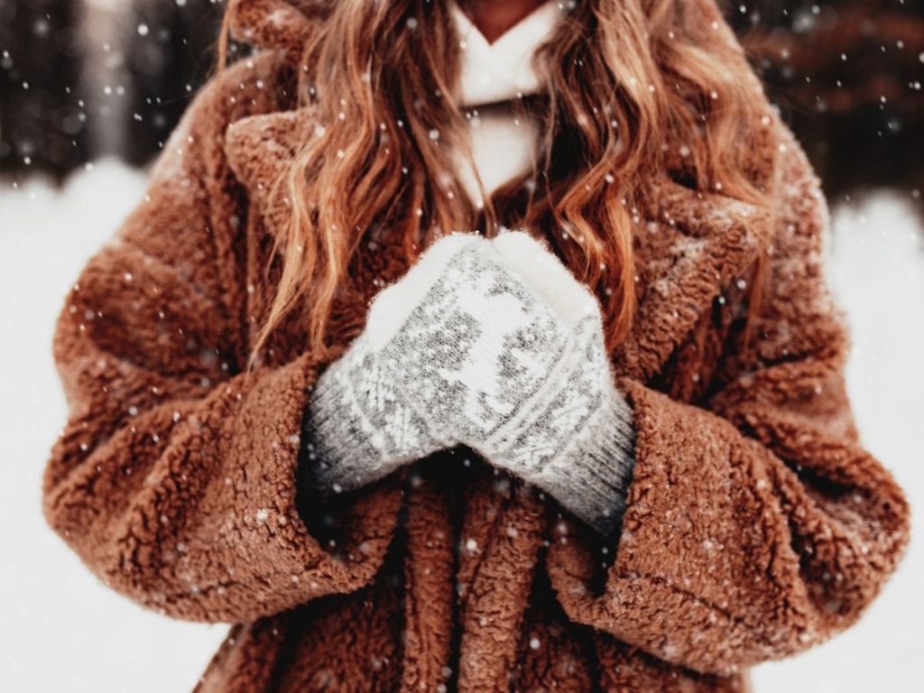 A person wearing a warm brown coat and gray patterned gloves holding their hands together in prayer during snowfall in winter.