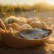 A woven wicker basket filled with bread rolls and two small fish sitting on a sandy beach under warm, golden sunset light.